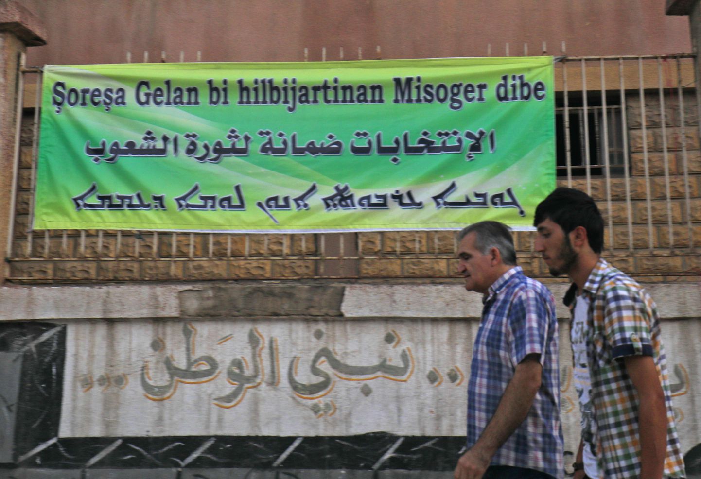 Valimakutsuvalt plakatilt Süüria kaguosas asuvas Qamishli linnas võib lugeda nii kurdi, araabia kui ka süüria keeles: «Valkimised on rahvaste revolutsiooni garant.»