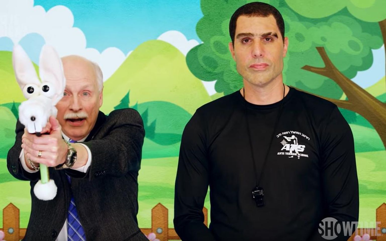 Sacha Baron Cohen (paremal) kolonel Erran Moradina koos relvakandmise õiguse pooldaja Philip Van Cleave’iga (vasakul) lastele tulirelvade kasutamist õpetamas.