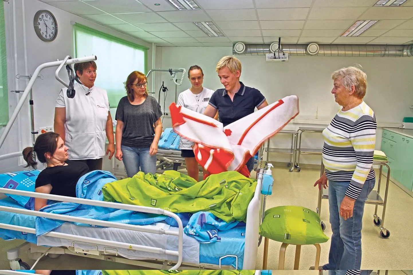 Õppeklassi voodis lamab hooldustöötaja õppekava õpilane Kristine Palm. Seisavad (vasakult) õppejõud Maire Aruots ning õpilased Ene Raba ja Liis Reitsnik. Tekiga jändab vabatahtlikku hooldustööd tegev Jana Tamm, kõrval on teine vabatahtlik Viia Konovalova.