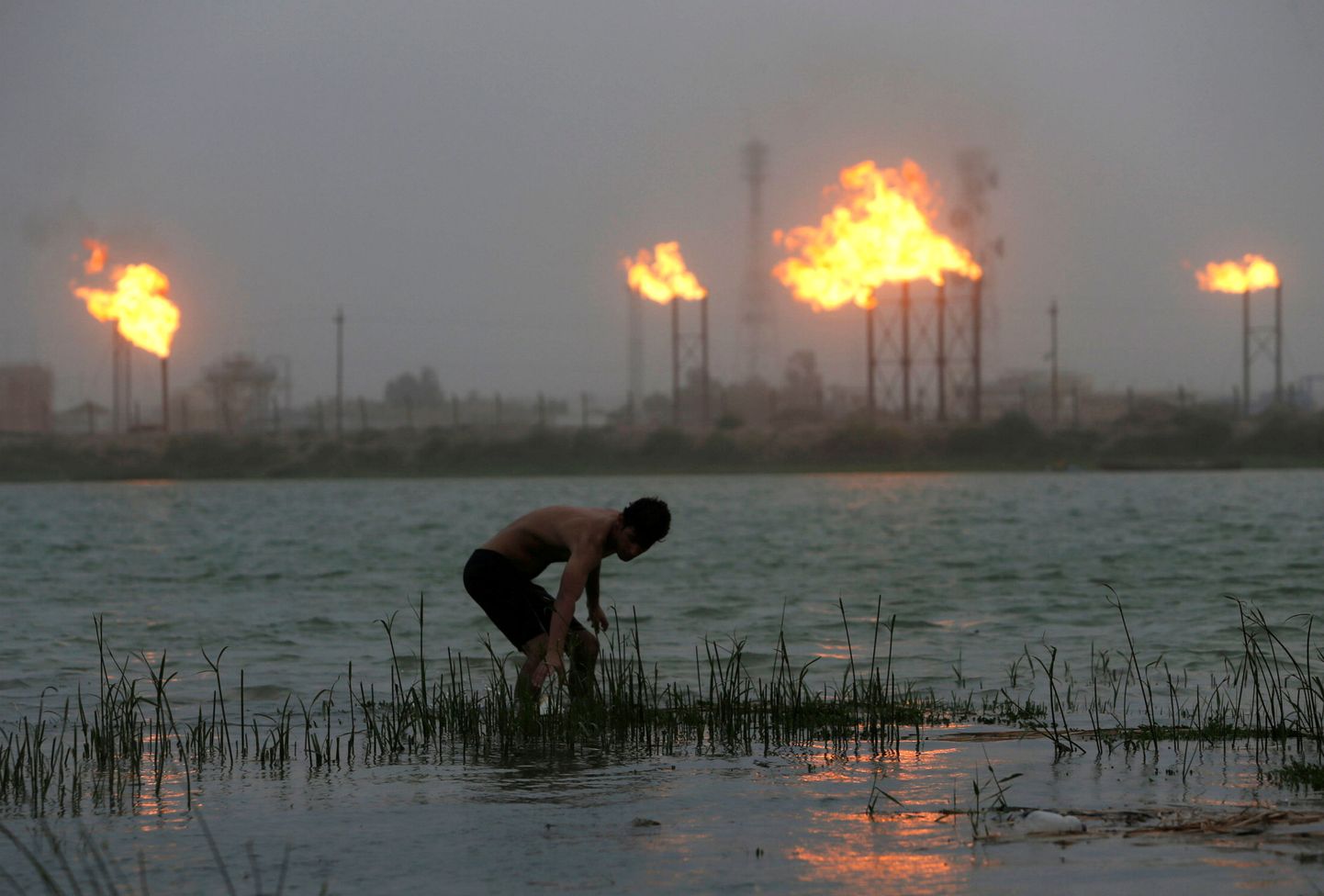 Mees seisab Basras Shaţţ al-'Arabi jões, taamal lõõmavad nafta rafineerimistehase korstnad.