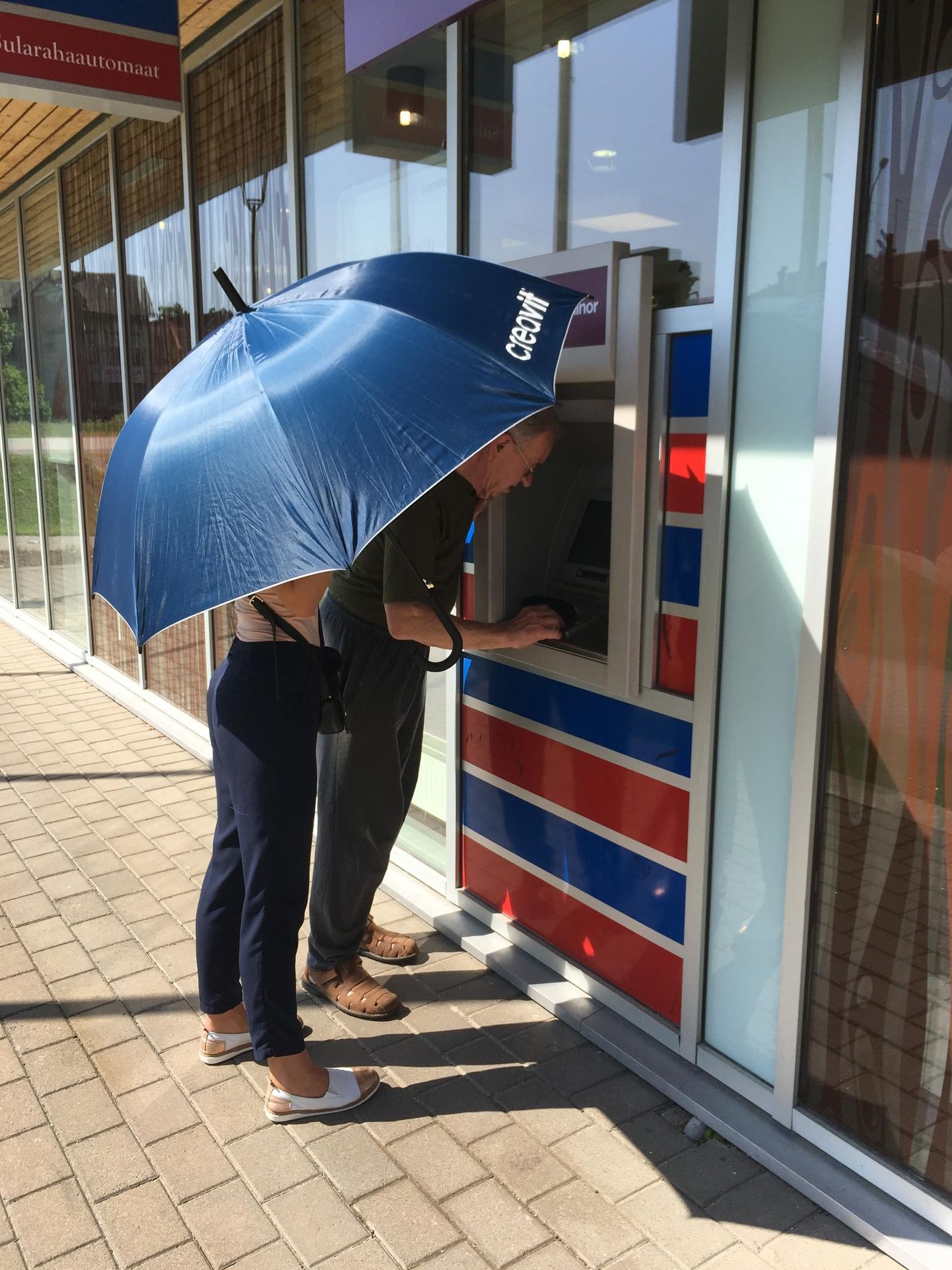 Kolmapäeva ennelõunal oli päike niivõrd ere, et pangatöötajal tuli vihmavarjuga automaadi kasutamiseks päikest varjata.