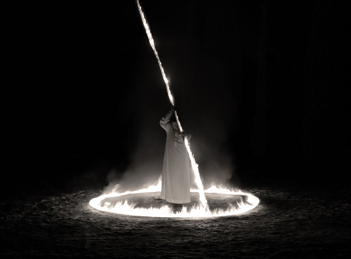 Художник, облаченный в белую мантию, стоит по одну сторону пространства, сформированного огненным световым кругом.