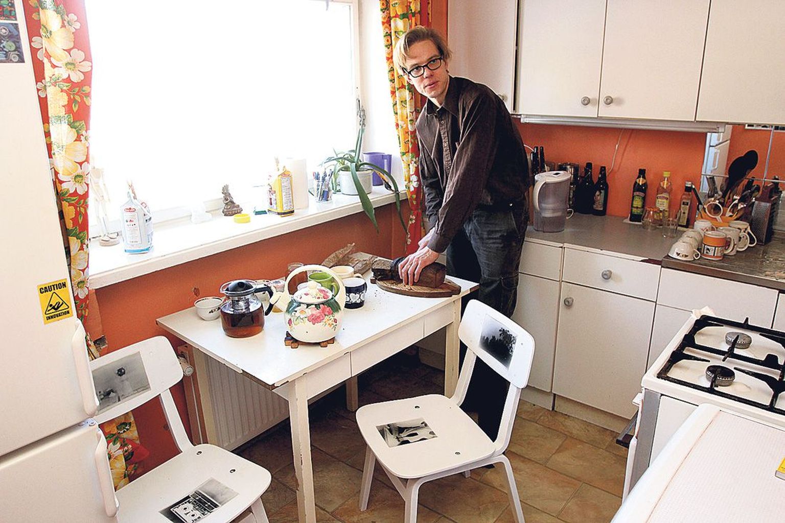 Indrek Grigor lõikas köögis lahti näituse avamiseks kingitud Muhu leiba. Laua kõrval on kaks Martiini fotodega tooli.