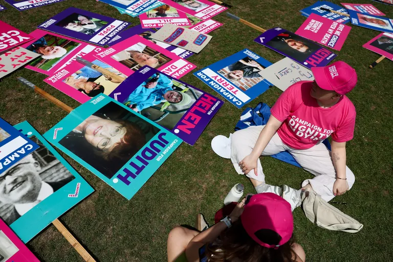 Asistētās pašnāvības legalizēšanas atbalstītājs nāvējoši slimiem cilvēkiem, ģērbies krekliņā un cepurē ar uzrakstu "Kampaņa par cieņpilnu nāvi" (Campaign for Dignity In Dying), sēž blakus uz zemes noliktam plakātam dienā, kad Lielbritānijas likumdevēji gatavojas balsot par likumprojektu, Londonā, Lielbritānijā, 2025. gada 20. jūnijā.
